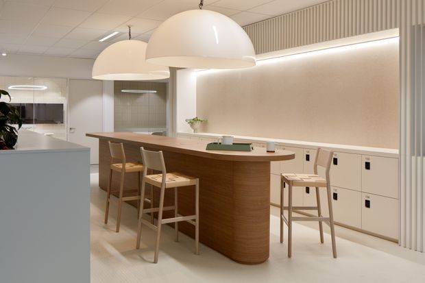 A bright, refined contemporary kitchen or communal kitchenette with a long curved timber island and integrated bar seating. Large white dome pendant lights hang above the island, while pale built-in cabinetry, a warm neutral feature wall, concealed strip lighting, and vertical ribbed detailing create a clean, softly illuminated interior. A small work area and cabinetry are visible in the background.