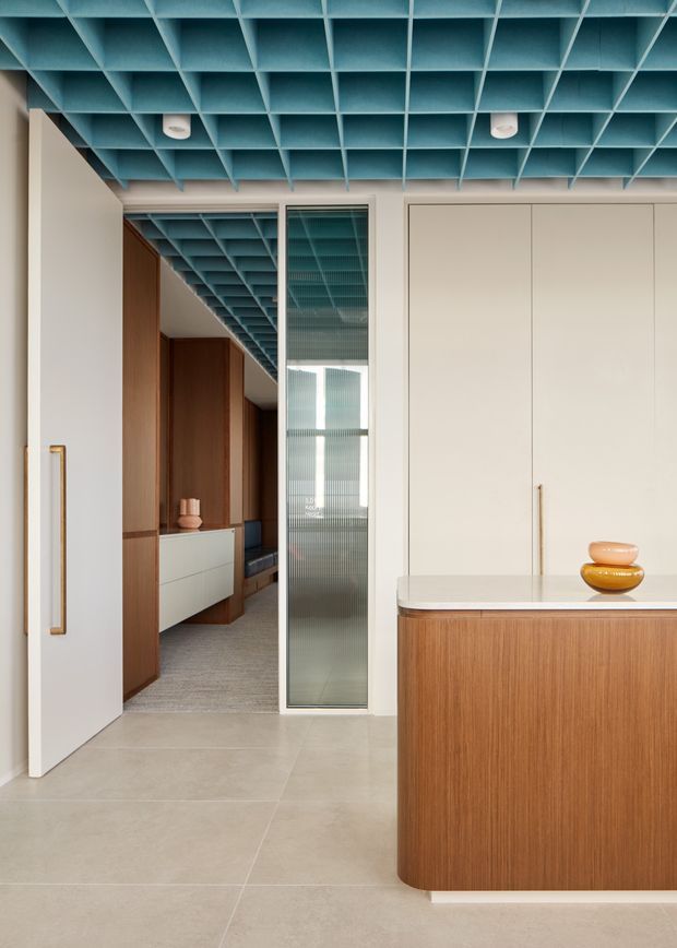 A refined contemporary interior featuring a cream-toned built-in kitchen or storage wall, a rounded timber island with a pale stone benchtop, and a corridor beyond with dark timber cabinetry. A distinctive turquoise-blue coffered ceiling grid spans the space, with a fluted translucent glass partition and warm brass-toned handles adding detail. The palette is warm and neutral with a strong blue ceiling accent.