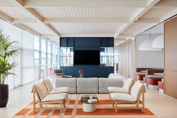 Bright open-plan living room with a large sectional sofa, two light timber lounge chairs, and a patterned area rug arranged around a low coffee table. Floor-to-ceiling glazing with sheer curtains lines the left side, while a dark blue built-in media wall anchors the room. The space extends to a dining or kitchen zone with stools and pendant lighting. A white painted timber-lined ceiling with exposed beams adds architectural character.