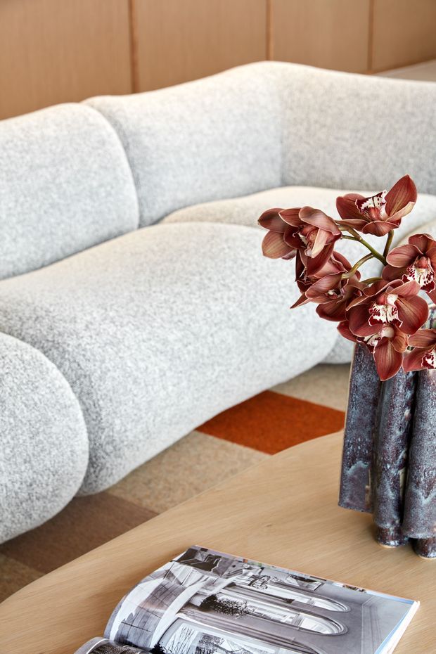 Close-up interior view of a softly furnished living room, dominated by a pale textured curved sofa, warm timber wall panelling, and a light timber coffee table. A dark mottled ceramic vase with burgundy orchid flowers sits on the table beside an open design magazine. Geometric terracotta and neutral floor tiles add a subtle accent beneath the sofa. The styling is refined, warm, and contemporary.