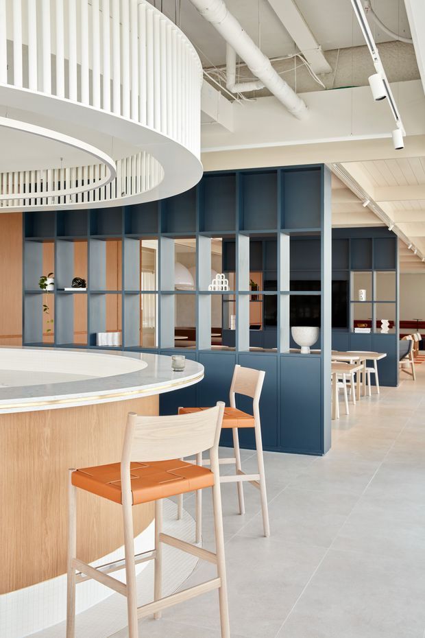 Bright contemporary commercial hospitality interior featuring a curved bar or reception counter with a light timber front and pale stone or solid-surface top. Deep blue fixed display shelving and glazed partition joinery divide the space, with warm timber accents, orange upholstered bar stools, white dining furniture, pale tiled flooring, and a prominent curved white slatted ceiling feature. Exposed services and suspended linear lighting contribute an open, modern fit-out.