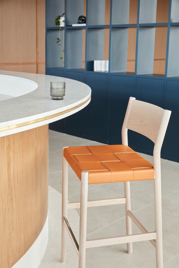 A close-up interior view of a refined contemporary kitchen or bar area. A curved pale stone island with a light timber base and thin brass-gold trim dominates the foreground, paired with a light timber bar stool featuring a woven orange leather seat. The background has full-height dark blue built-in cabinetry and open shelving with timber backing, creating a warm contrast with the pale tiled floor and natural light.