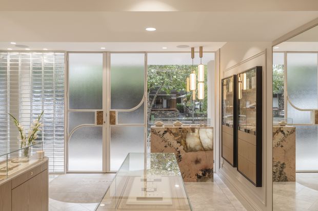 Bright, refined contemporary interior showing a compact open-plan living and dining area with a glass-topped table, timber cabinetry, decorative framed wall panels, and a sculptural stone island or counter. Frosted and clear glazed partitions with curved detailing open toward a landscaped courtyard, while warm pendant lighting and pale finishes create an elegant, airy atmosphere.