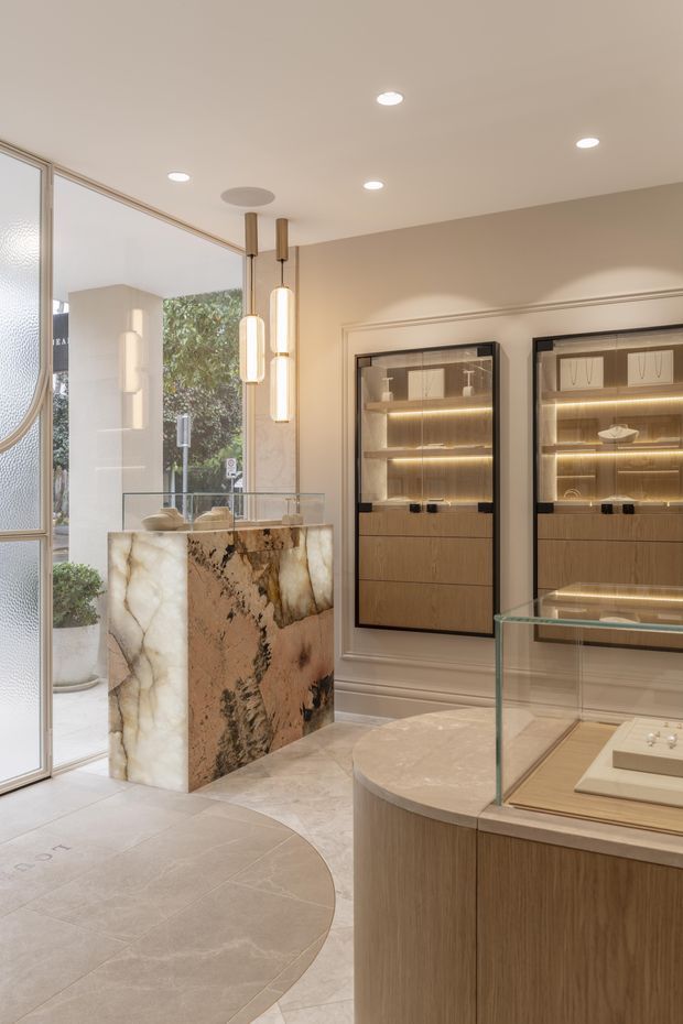 Elegant retail or showroom interior with warm neutral finishes, illuminated glass display cabinets, a curved timber display counter with glass top, and a dramatic natural stone or onyx reception/display island. Full-height glazing opens toward greenery outside. Decorative pendant lights and recessed ceiling lighting create a refined, softly lit atmosphere.