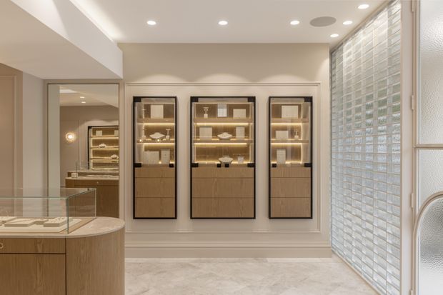 A refined retail or showroom interior with three illuminated glass-front display cabinets set into a decorative paneled wall. Warm wood cabinetry and a curved glass-topped display counter occupy the foreground, while large translucent glass-block glazing on the right admits soft daylight. The space has recessed ceiling lighting, pale stone-look flooring, and a warm neutral palette.