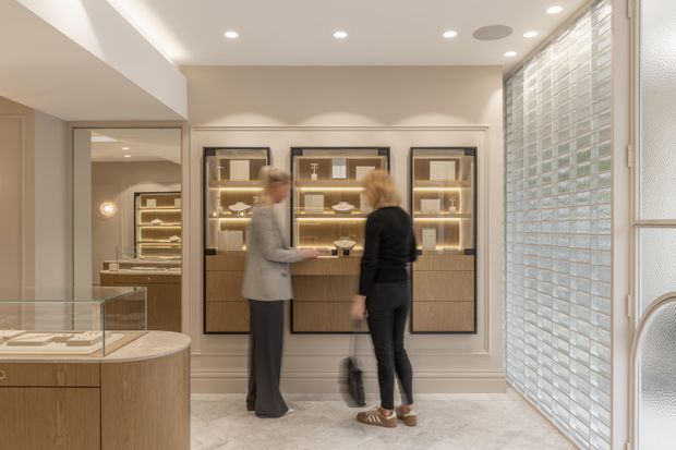 A refined retail showroom or jewellery display interior with illuminated glass-fronted cabinetry, warm wood joinery, pale stone flooring, and a curved glass display counter. Two people stand near the central display wall. The space has recessed ceiling lighting, decorative wall moulding, and a textured glass-block window or partition along the right side, creating a bright, softly luxurious atmosphere.