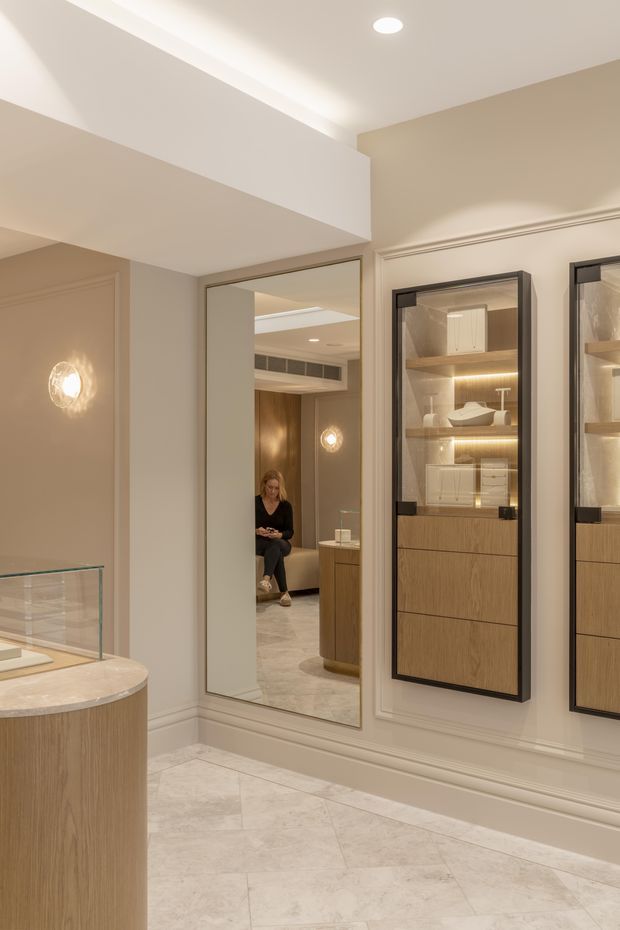A refined, softly lit interior showroom or retail display space with warm beige walls, decorative wall mouldings, pale stone floor tiles, and integrated illuminated display cabinets containing ceramics and small objects. A large mirror reflects a seated person and additional cabinetry, while a rounded timber display counter with a light stone top occupies the foreground. Recessed ceiling lighting and decorative wall sconces create a warm, luxurious atmosphere.