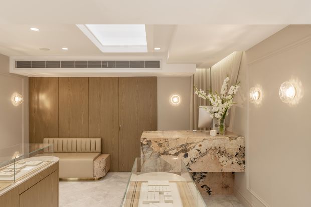 A refined, softly lit luxury retail showroom or display space with a marble reception counter, glass-topped display cases, warm timber cabinetry, upholstered bench seating, decorative wall sconces, and a recessed skylight. The neutral palette and layered lighting create an elegant, high-end atmosphere.