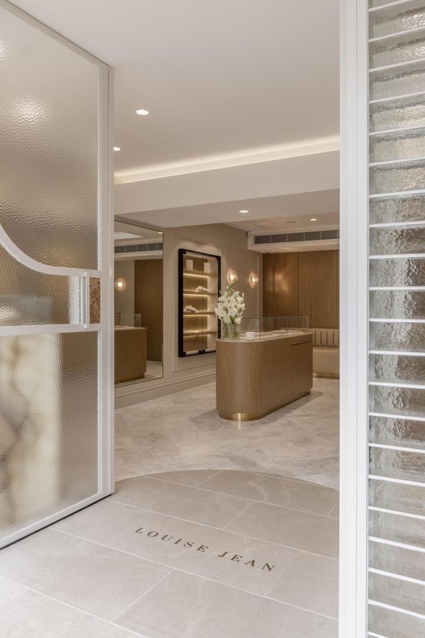 A refined luxury retail or reception entry viewed through frosted glass doors. The space features a curved timber reception counter, warm wood wall panelling, illuminated display cabinetry, upholstered bench seating, pale large-format stone-look flooring, mirrored surfaces, and discreet recessed lighting. The palette is warm neutral with cream, beige, and timber tones.
