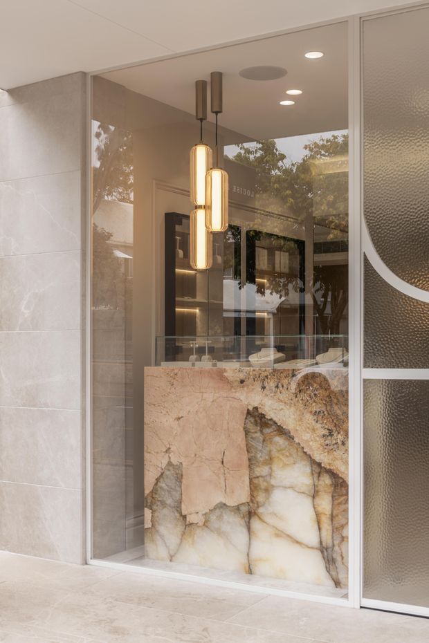 A refined commercial interior viewed through a glazed storefront, featuring a dramatic translucent stone reception or display counter with warm amber veining, pale stone wall and floor finishes, dark built-in shelving, and a cluster of elongated illuminated pendant lights. Textured glass and curved metal detailing frame the entrance, while reflections reveal trees and the surrounding exterior.