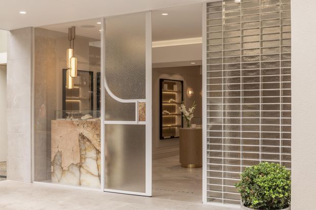 A refined residential entry viewed from outside, featuring a partially open decorative glass pivot door, a translucent textured-glass side panel, a pale stone-clad entry volume, and a striking natural stone feature panel with warm veining. The entry opens toward a softly lit interior with timber-toned flooring, a curved wood console, display shelving, and warm pendant lighting. A gridded light brick or tile screen frames the right side, with a clipped potted shrub in the foreground. The palette is warm, neutral, and luxurious with layered stone, glass, metal, and timber finishes.