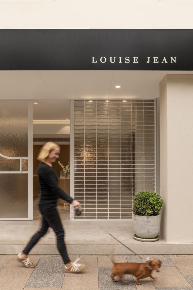 Street-level view of a refined commercial storefront with a black fascia sign reading “LOUISE JEAN.” The entry features textured and frosted glass doors, a large glazed tile or brick-patterned screen, recessed ceiling lighting, pale stone paving, and a potted clipped shrub. A pedestrian and dog are passing in the foreground.