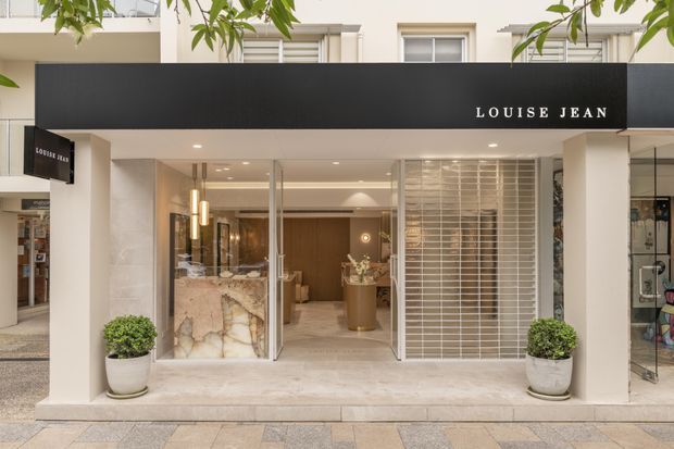 Street-level view of a refined boutique retail storefront with a black projecting fascia sign reading “LOUISE JEAN.” The entrance is framed by pale stone or plaster surfaces, full-height glazing, a white metal security grille or screen, and symmetrical potted topiary plants. Warm interior lighting reveals a polished stone reception or display counter and restrained luxury finishes inside.