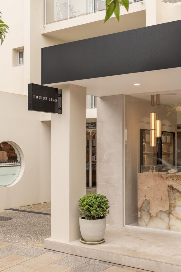 A refined street-facing boutique storefront with a pale rendered facade, large dark charcoal projecting fascia, and a black hanging sign reading “LOUISE JEAN.” The entrance is framed by light stone-look wall cladding and a dramatic natural stone reception or display counter visible inside, illuminated by elongated pendant lights. A potted topiary sits beside the entry, while neighbouring retail details and a rounded display window are visible to the side.