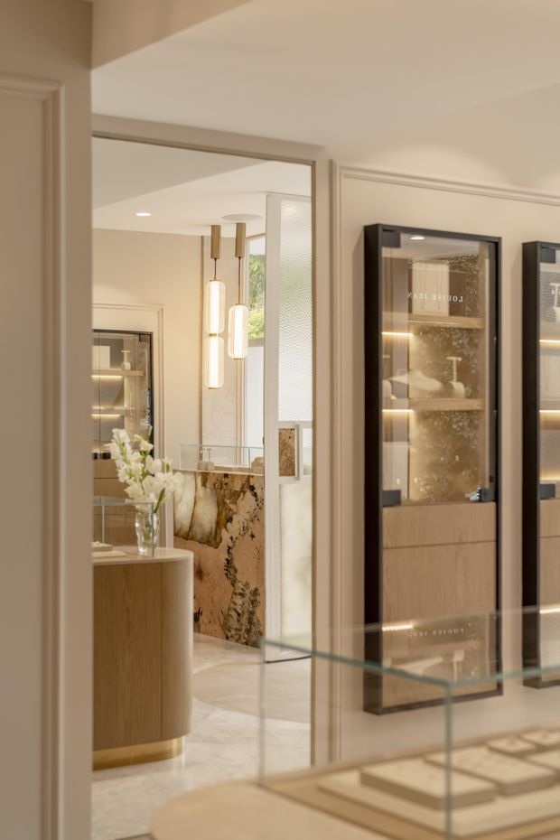 A refined interior view showing a warm, luxurious display and dining area. A large wall mirror reflects a stone island or counter, pendant lights, glass shelving, and cabinetry. The foreground includes a curved timber pedestal, a glass-topped surface, and illuminated black-framed display cabinets with wood drawers. Decorative wall mouldings, pale stone flooring, brushed metallic details, and soft beige tones create an elegant contemporary-classic atmosphere.