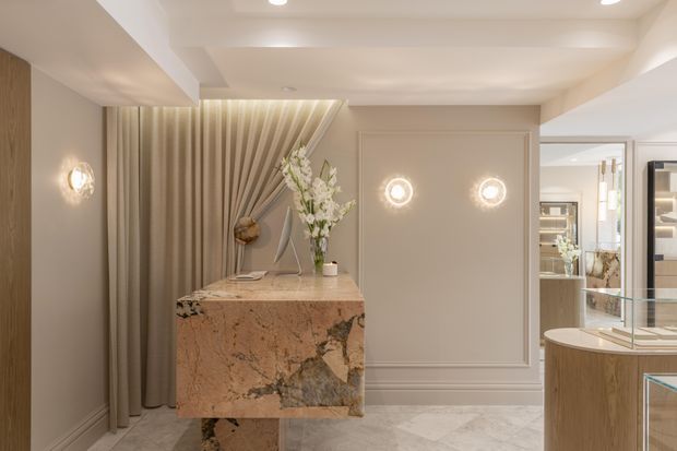 A refined, warmly lit interior featuring a sculptural stone console or island with a matching pedestal base, set against taupe decorative wall panelling and full-height pleated curtains. Marble or natural stone flooring, mirrored and glass display joinery, recessed ceiling lighting, and decorative wall sconces create a luxurious showroom-like space. Floral arrangements and display objects are present, but the dominant features are the fixed finishes, stone surfaces, and built-in cabinetry.