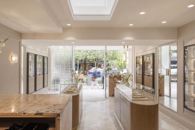 Bright contemporary showroom or retail interior with a central aisle, large marble-topped display counters, curved timber cabinetry, glass-fronted wall display units, pale herringbone flooring, recessed ceiling lights, and a large skylight. Full-height glazing opens toward an outdoor courtyard or entrance area. The space appears refined, warm, and highly polished.