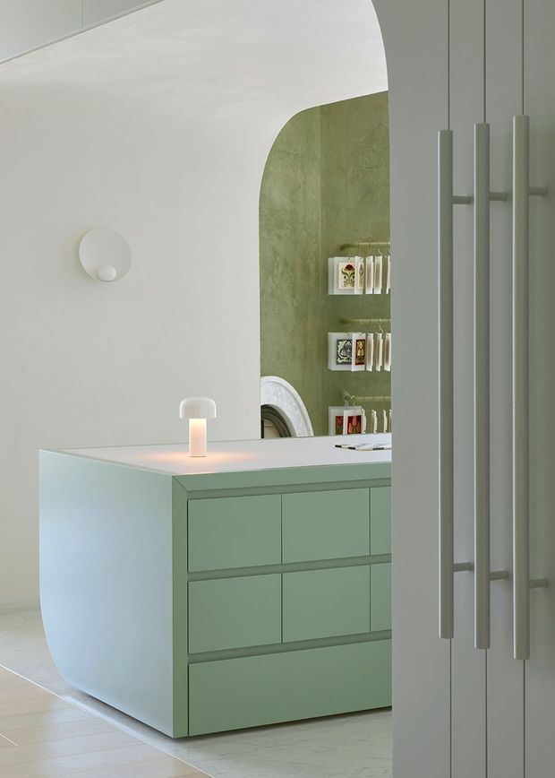 A softly lit contemporary interior featuring a pale sage-green built-in drawer cabinet with a rounded white end panel and integrated countertop. Behind it is a large arched opening with textured green wall treatment, display shelving, and a partial mirror or artwork. Tall pale cabinetry with long vertical handles occupies the right foreground, while minimalist pendant and wall lighting reinforce the calm, sculptural design.