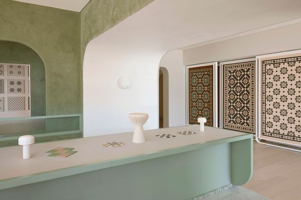 A refined, pastel-toned interior featuring a long mint-green display counter or island with rounded ends, white arched openings, and decorative geometric patterned panels displayed along the rear wall. Small sculptural lamps and tile-like samples sit on the counter, creating a curated showroom or design studio atmosphere.