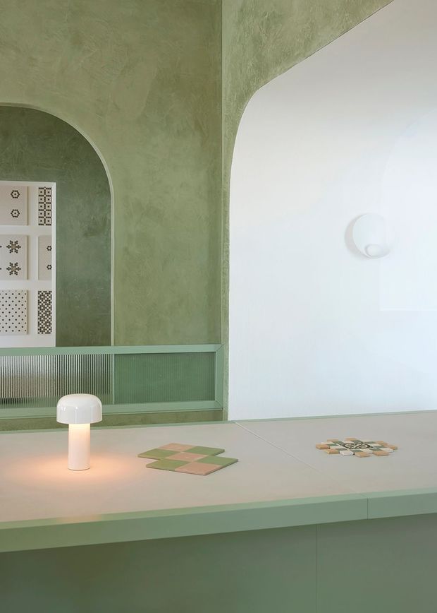 A softly lit contemporary interior display featuring a pale green textured plaster wall with a large rounded arch and curved mirror or opening, a ribbed glass-fronted built-in counter, and a pale green worktop or island. Small tile samples and a glowing white table lamp sit on the surface, while a white wall with a minimalist circular light fixture occupies the right side. The palette is muted sage green, pale pink, cream and white, with rounded geometric detailing throughout.
