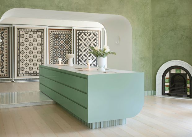 A distinctive kitchen with a large pale green island featuring rounded corners, integrated drawer cabinetry, and a ribbed metal plinth. The room has a mottled sage-green wall finish, pale timber flooring, a curved white archway, patterned decorative panels at the rear, and a small arched fireplace with a dark grate. A vase of flowers and countertop accessories sit on the island.