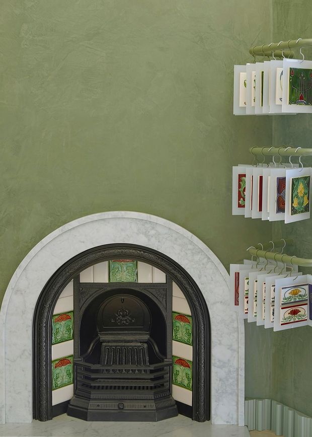 A close-up interior view of a decorative period fireplace set into a green textured plaster wall. The fireplace has an ornate dark metal surround, a pale marble arched mantel and colourful patterned tiles. Small illustrated cards or prints are displayed on wire racks to the right, with a light-toned floor and radiator visible at the base.