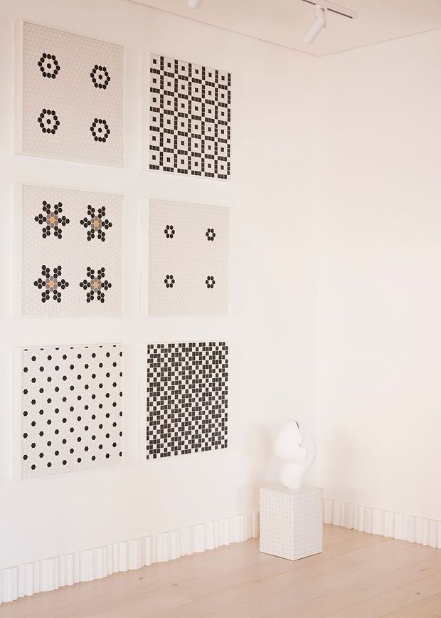Bright minimalist gallery-like interior with white walls, pale timber flooring, decorative monochrome geometric artworks arranged in a grid, and a white sculptural object displayed on a gridded pedestal. Track lighting and fluted white skirting add subtle architectural detail.