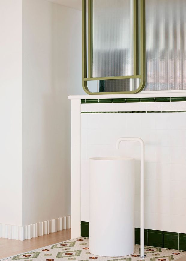 A bright, close-up view of a refined bathroom corner featuring white tiled walls with dark green trim, a patterned geometric mosaic floor, a sculptural white cylindrical basin or pedestal fixture, and an olive-green framed reeded-glass window or mirror. The space has soft white walls, rounded detailing, and a clean contemporary aesthetic.