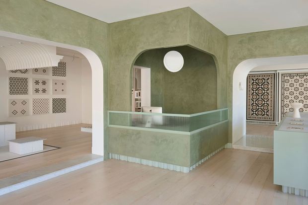 A refined gallery or showroom interior with pale timber flooring, textured sage-green plaster walls, rounded architectural openings, and a central glazed reception or display counter. Decorative patterned tiles are exhibited on the walls, while white built-in cabinetry and softly curved detailing create a calm, contemporary setting.