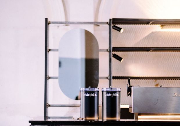 Close-up view of a refined café or commercial service counter featuring black cylindrical canisters, a metallic countertop appliance, black open shelving with integrated spotlights, and a large rounded-arch mirror or reflective panel. The space has a pale plaster-like backdrop with curved architectural detailing and a high-contrast black, silver, and warm-white palette.