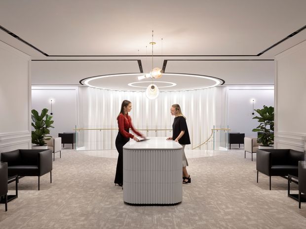 A refined contemporary reception lobby with a central rounded, vertically fluted reception desk, two staff members, plush patterned carpet, black lounge chairs, indoor plants, sheer curtains, and a striking circular layered pendant light. White paneled walls, concealed linear ceiling lighting, and a glass balustrade create a bright, symmetrical and luxurious hospitality or commercial interior.