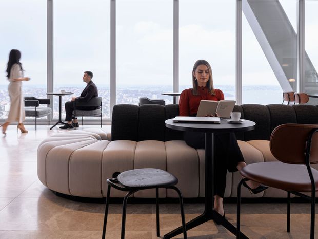 A sophisticated high-rise lounge or hospitality interior with floor-to-ceiling glazing overlooking a distant cityscape. Curved upholstered modular seating in cream and charcoal surrounds small round black tables, while contemporary lounge chairs and a marble-topped stool complete the refined setting. Several people are using the space for seating and laptop work. The interior has a bright, polished, modern appearance with exposed structural glazing supports.