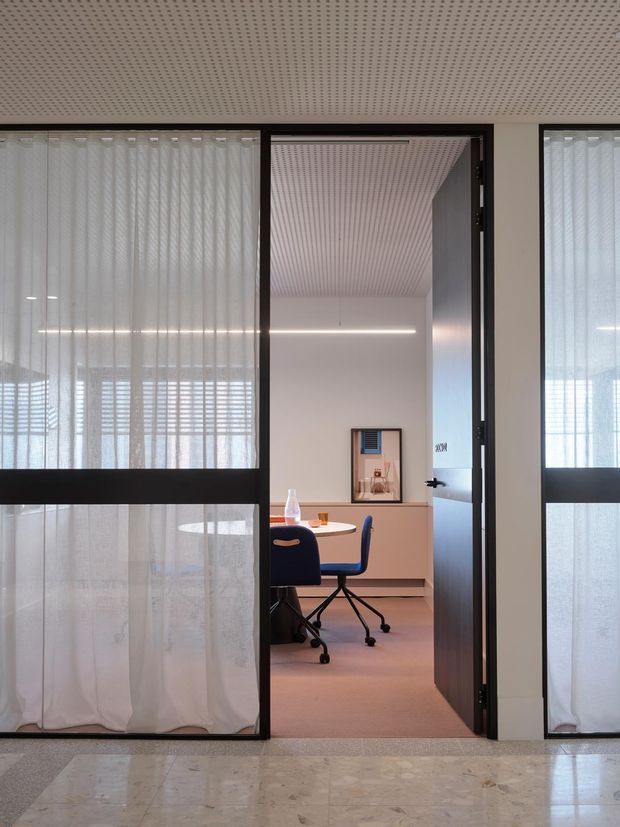 A contemporary office interior viewed through black-framed glazed partitions and an open glass door. Sheer white curtains filter daylight on both sides, while a compact meeting or work area beyond contains a round table, blue office chairs, a pale built-in credenza, and a framed wall artwork. The space has a perforated acoustic ceiling, recessed linear lighting, and a polished light stone or terrazzo-like floor in the foreground.