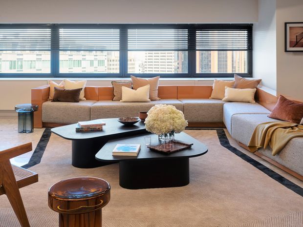 A bright, spacious contemporary living room with a large L-shaped sectional sofa beneath expansive horizontal windows with venetian blinds. Two black sculptural coffee tables sit on a large neutral area rug, accompanied by layered cushions, a leather stool, timber seating, and warm decorative accents. Urban apartment buildings are visible through the windows.
