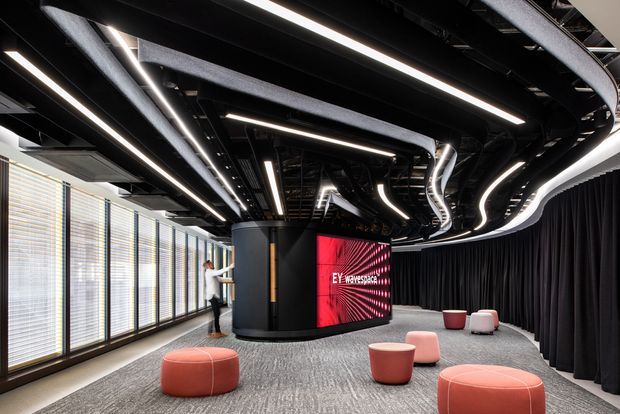 A dramatic contemporary commercial interior with an open presentation or collaboration area. The space features a large curved digital display, exposed black ceiling services, extensive linear and sinuous integrated LED lighting, full-height windows with horizontal blinds, dark curtains, grey carpet, and scattered rounded pink and coral upholstered ottomans.