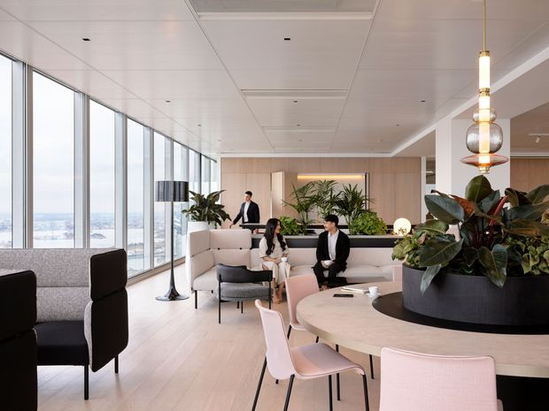 A bright, sophisticated high-rise communal lounge with floor-to-ceiling glazing, pale timber flooring, built-in upholstered seating, indoor planting, a large round dining or meeting table, and pendant lighting. The space overlooks an urban landscape and includes several people, but the focus is the refined shared interior.
