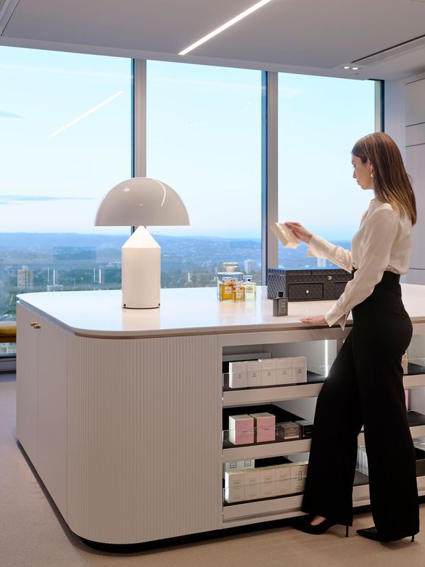 A contemporary high-rise interior featuring a refined beauty or retail-style island counter positioned in front of floor-to-ceiling windows. The curved white island has ribbed cabinetry, open display shelving, and a glossy white benchtop, with a sculptural dome table lamp and neatly arranged product boxes. A woman stands beside the counter, while expansive glazing provides a panoramic urban landscape view. Linear integrated ceiling lighting and minimalist detailing create a polished, luxurious atmosphere.