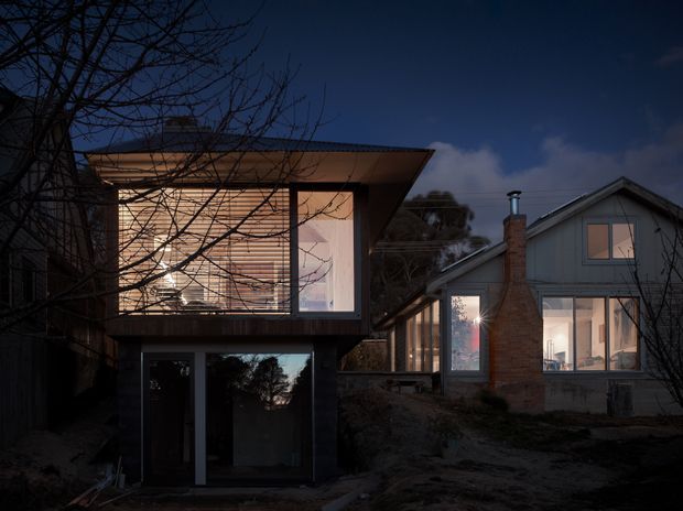 Twilight exterior view of a residential property comprising a dark, box-like contemporary extension in the foreground and a pitched-roof house behind it. The extension features large illuminated glazing, horizontal timber blinds, dark cladding, and a broad projecting flat roof. The existing house has light vertical cladding, a brick chimney, and large windows. Leafless trees and a sloping garden surround the buildings.