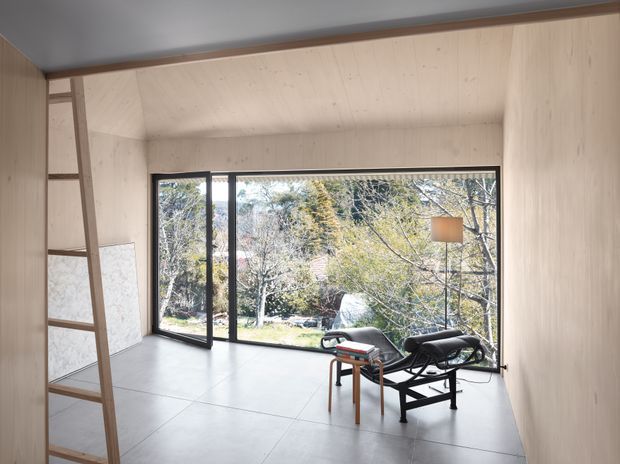 A compact, minimalist interior room clad in pale timber panels, with a large black-framed glazed opening overlooking trees and a garden. A simple black lounge chair, small side table, and leaning ladder occupy the otherwise open space. The room has a smooth light grey tiled floor, a low plain ceiling, and abundant natural light.