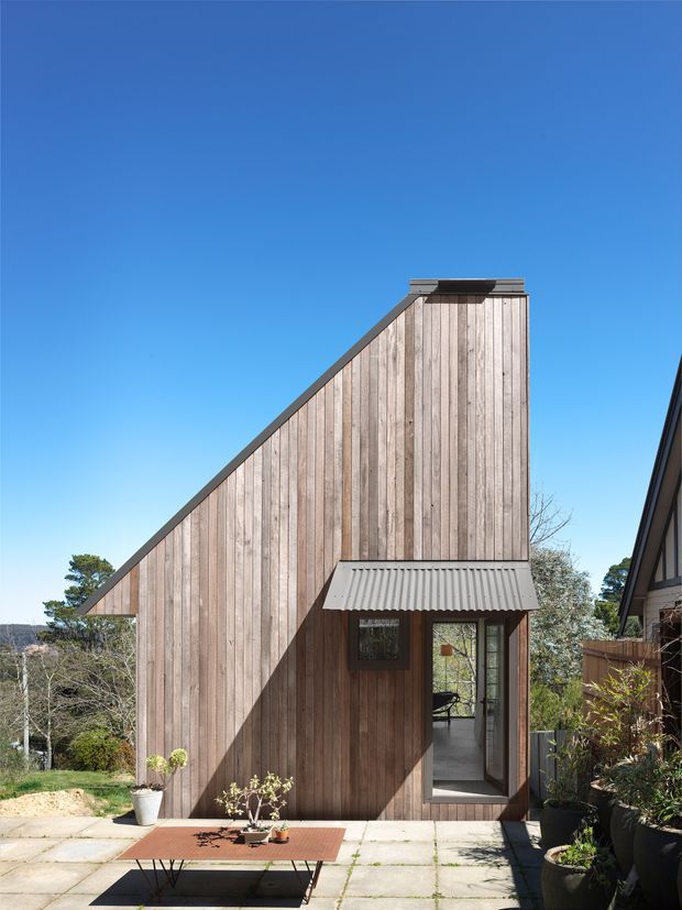 Exterior view of a compact, sharply geometric building clad in vertical weathered timber boards. The facade has a dramatic sloping roofline rising to a flat-topped high point, dark trim, and a small corrugated metal awning over a glazed entry door. It sits beside a paved outdoor area with potted plants, under a clear blue sky.