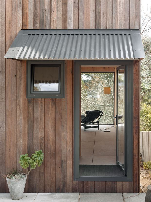 Close-up exterior view of a compact timber-clad building entrance. Vertical weathered wood boards surround a dark grey corrugated metal awning and matching dark-framed window and glazed door. The open doorway reveals a simple interior with a lounge chair, pendant light and large glazing looking toward trees. A concrete threshold and potted plant sit in the foreground.