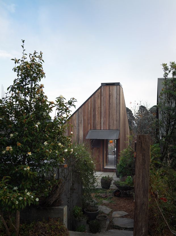 A narrow landscaped garden path leads toward a compact timber-clad building with a distinctive steeply sloping, asymmetrical gabled facade. Vertical weathered wood boards, dark roof trim, a small corrugated metal awning, and glazed entry door are visible, with dense planting and timber fencing framing the approach.