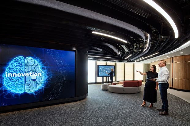 A contemporary corporate innovation and presentation space with a dramatic black ribbed ceiling, integrated curved lighting, dark carpet flooring, timber wall panelling, and a large digital display featuring an innovation graphic. Curved modular lounge seating and presentation screens create an open, high-tech workplace or showroom environment.