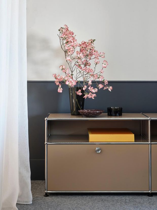 A refined interior vignette featuring a low modern sideboard against a two-tone wall. The cabinet has a warm taupe-brown drawer front, metallic tubular framing, and an open shelf containing a mustard-yellow box. A tall dark vase with delicate pink blossoms, a shallow decorative bowl, and a black vessel sit on top. White curtains frame the scene, with grey carpet underfoot.