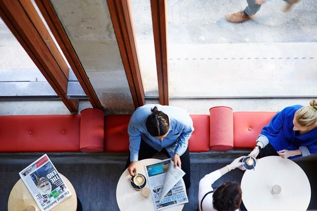 Overhead view of a modern café or commercial lounge with red upholstered banquette seating, round white tables, large timber-framed glazing, and patrons reading newspapers and drinking coffee. The space has a polished, lively urban hospitality atmosphere.