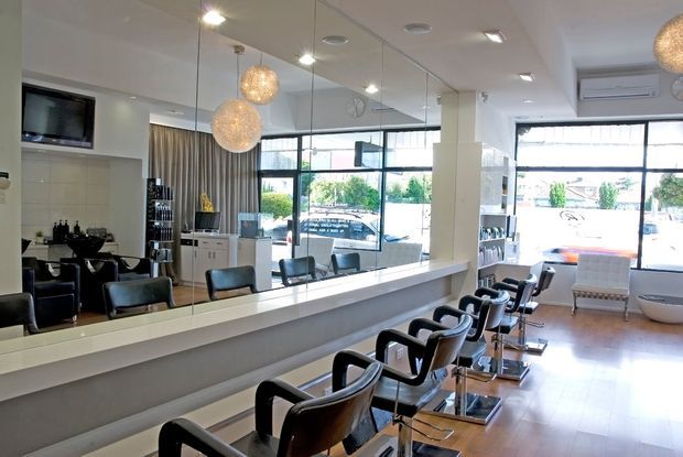 Bright contemporary hair salon interior with a long styling counter and multiple black salon chairs facing a large mirrored wall. The space features pale flooring, white walls and ceiling, extensive glazing, recessed lighting, and spherical pendant lights, with a waiting area and salon equipment visible toward the rear.