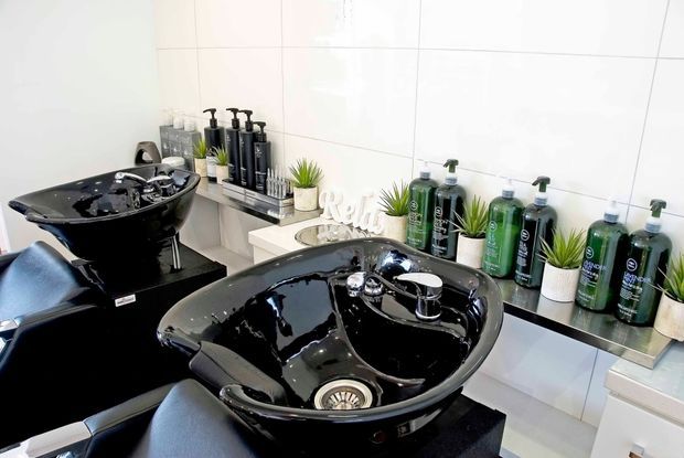 Bright commercial hair salon shampoo area with two glossy black ceramic wash basins, padded black salon chairs, wall-mounted shelving, salon product bottles, small potted plants, and white tiled walls.