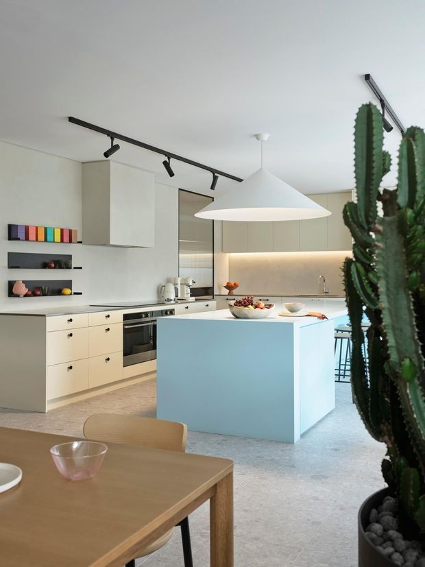 Bright contemporary open-plan kitchen and dining area with streamlined pale cabinetry, integrated appliances, a large light-blue island, pale stone-look flooring, and a sculptural white pendant light. Black track lighting, open display shelves, and tall potted cacti add graphic contrast and a modern, refined character.
