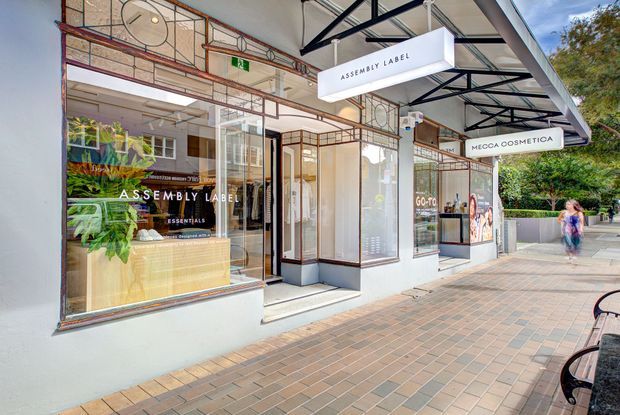 Street-level exterior view of a contemporary retail storefront in a commercial precinct. The facade features large shopfront glazing, white rendered walls, dark metal framing, exposed structural steel awnings, and prominent hanging signage for Assembly Label and Mecca Cosmetica. A paved pedestrian walkway and landscaped greenery are visible outside.