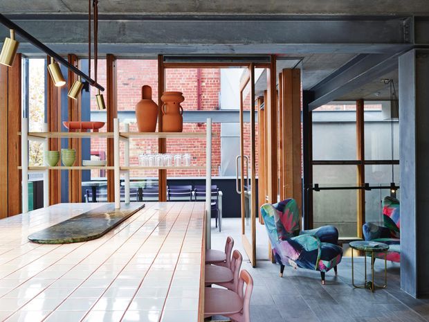 Open-plan contemporary interior combining a large tiled kitchen island with integrated shelving, a dining area, and a colourful lounge nook. Exposed concrete ceiling beams and columns contrast with warm timber-framed glazing, red brick walls, dark flooring, and vivid upholstered armchairs. The kitchen opens visually toward a dining or living space, with pendant lighting and strong indoor-outdoor connections.